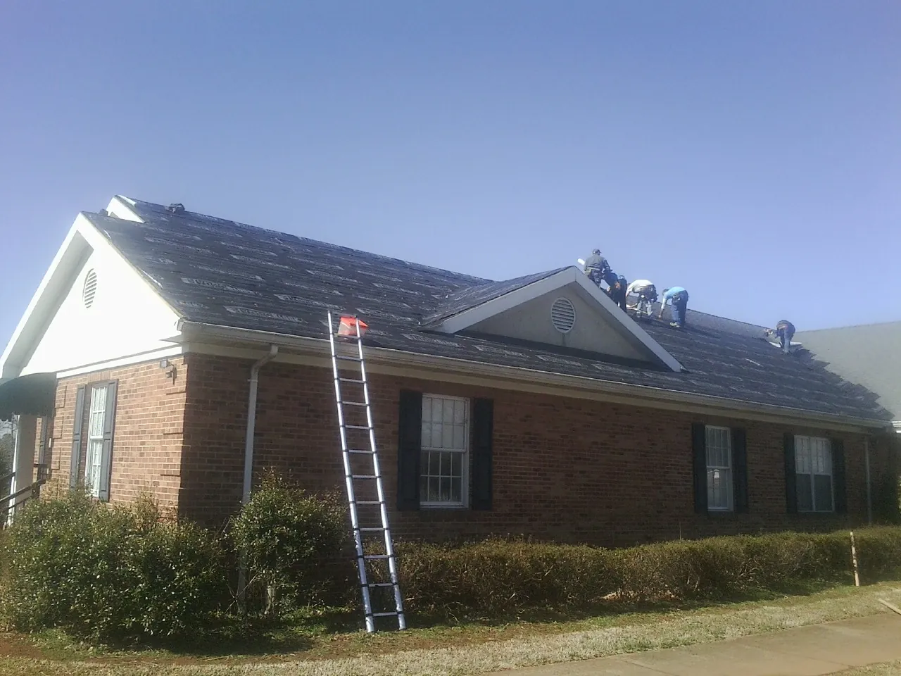Crew tearing shingles off a brick ranch roof with a ladder set — Emergency Roof Repair work by our crew in Sand Lake