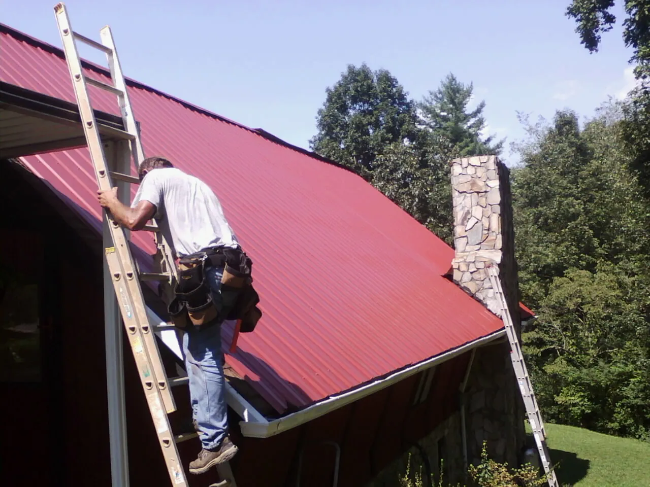 Roofer on a ladder beside a red metal roof and stone chimney — Soffit & Fascia Repair by Roofing Brothers in Sand Lake